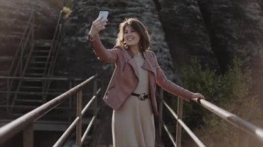 Ağır çekim. Yakın görüş. Dağlarda köprü boyunca yürüyen güzel bir kadın. Cep telefonundan selfie çekiyor. Omuz hizasında koyu renk saçlarını düzeltiyor. Sakson İsviçre Ulusal Parkı