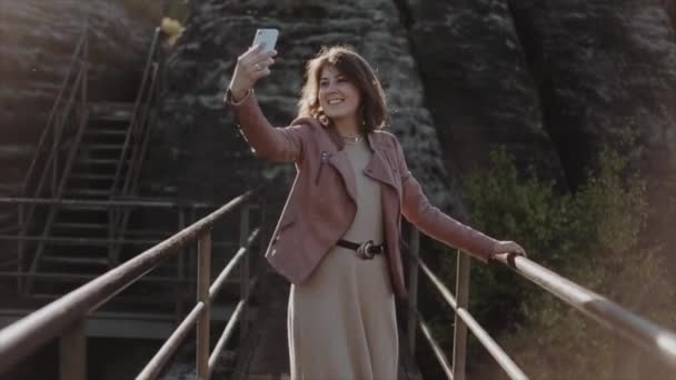 En cámara lenta. Vista cercana. Hermosa mujer caminando a lo largo del puente en las montañas. Haciendo selfies en su móvil. Arreglándose el pelo oscuro de su hombro. Parque Nacional Sajón Suiza — Vídeos de Stock
