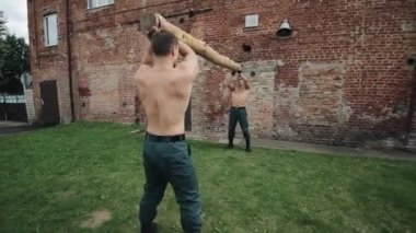 İki kaslı üstsüz adam bir kütüğü kaldırarak ve onu bir omzundan diğerine koyarak birlikte çalışıyor. Kamera sağdan sola hareket ediyor