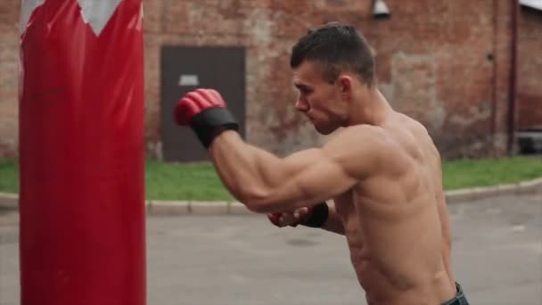 Vista lateral cercana del boxeador masculino muscular practicando golpes y patadas con un saco de boxeo al aire libre — Vídeo de stock