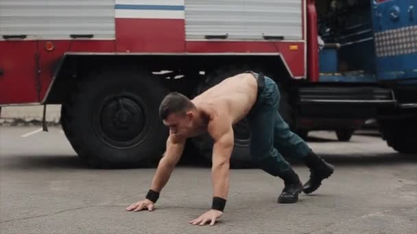 En cámara lenta. Atractivo joven atleta sin camisa ejercitando flexiones con saltos. Gran camión de bomberos en el fondo. Vista lateral — Vídeos de Stock