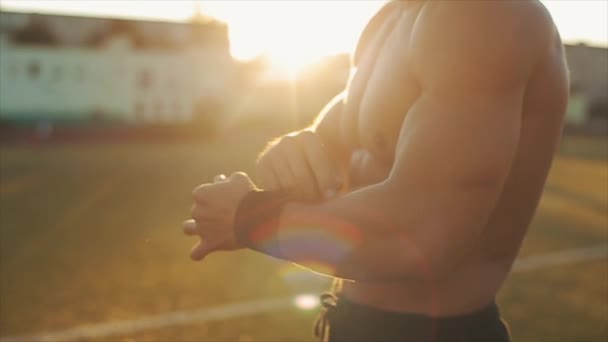 Vista lateral de perto de um desportista muscular colocando em apoio de pulso preto em mãos. Sol brilhante no fundo — Vídeo de Stock