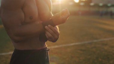 Siyah bilek desteği takan kaslı bir sporcunun yakın görüntüsü. Arkaplanda parlak güneş