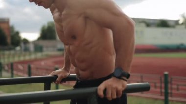 Spor sahasında vücut ağırlığı şınavı yapan üstsüz, kaslı bir sporcunun yakın çekim görüntüsü. Arka planda futbol oynayan insanların bulanık görüntüsü