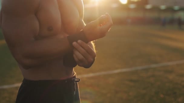 Vista lateral cercana de un deportista musculoso que se pone el soporte de muñeca negro en las manos. Sol brillante en el fondo — Vídeos de Stock