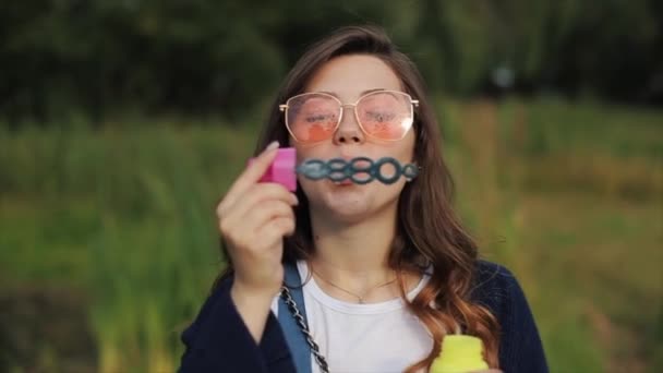 Mooie jonge vrouw in een zonnebril blaast zeepbellen buiten en draait rond. Goed zicht. Langzame beweging. meisje in trendy oranje zonnebril — Stockvideo