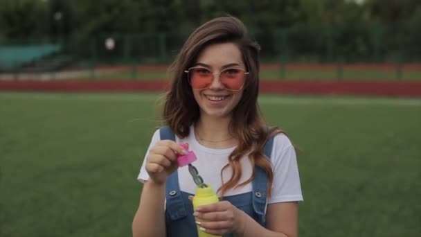 Schöne junge Frau mit Sonnenbrille, die draußen Seifenblasen pustet und sich herumdreht. Nahaufnahme. Zeitlupe — Stockvideo