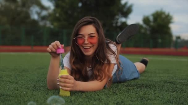 Hermosa mujer joven en gafas de sol soplando burbujas de jabón al aire libre y girando alrededor. Vista cercana. En cámara lenta. la chica se ríe — Vídeos de Stock