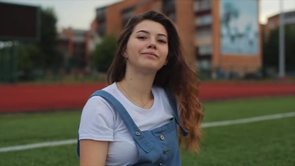 Porträtt av vacker brunett kvinna inblue solklänning sitter på gräsmattan nära ett fotbollsmål på stadion. solljus. En flicka med en perfekt figur och i bra form. vacker flicka i orange — Stockvideo