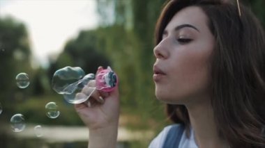 Güneş gözlüklü genç bir kadın dışarıda sabun köpüğü üflüyor ve etrafında dönüyor. Yakın görüş. Yavaş çekim