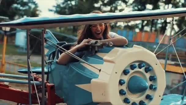 Bela menina sorridente em óculos de sol montando um carrossel de crianças. Vista próxima. Movimento lento. menina bonita andar de avião carrossel — Vídeo de Stock