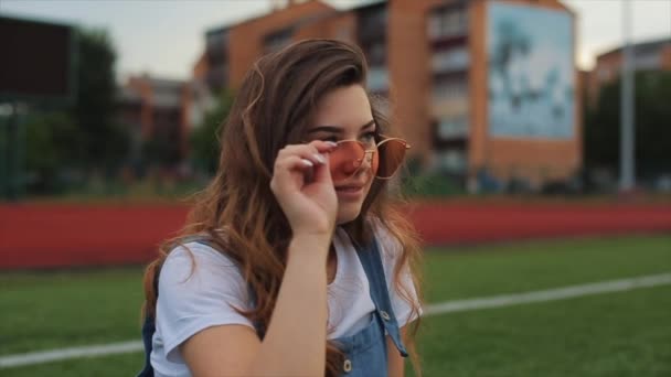 Portrét krásné brunetky žena inblue Sundress sedí na trávníku v blízkosti fotbalového cíle na stadionu. sluneční svit. Dívka s dokonalou postavou a ve skvělé formě. hezká dívka v oranžové — Stock video