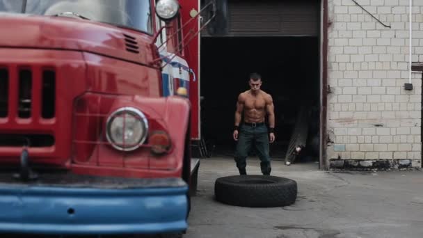 Joven guapo sosteniendo un neumático y haciendo sentadillas cerca del camión de bomberos — Vídeo de stock