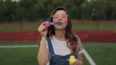 Güneş gözlüklü genç bir kadın dışarıda sabun köpüğü üflüyor ve etrafında dönüyor. Yakın görüş. Yavaş çekim