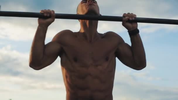 Guapo culturista haciendo pull-ups en la barra horizontal en el gimnasio exterior. Vista cercana. Hermoso amanecer en el fondo . — Vídeos de Stock