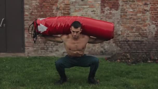 Un hombre guapo en cuclillas con un gran saco de boxeo en los hombros. Pared de ladrillo rojo en el fondo — Vídeos de Stock