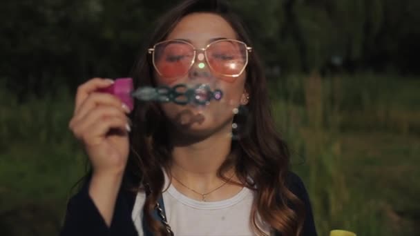 Bella giovane donna con gli occhiali da sole che soffia bolle di sapone all'aperto e gira intorno. Vista da vicino. Rallentatore — Video Stock