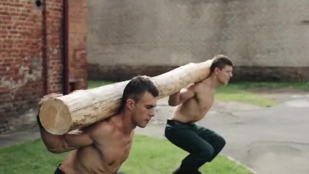 Vista cercana de dos hombres jóvenes en forma haciendo sentadillas con saltar sosteniendo un tronco sobre sus hombros. Entrenando afuera. Cámara moviéndose de arriba a abajo — Vídeos de Stock