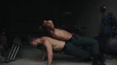 Sırtında başka bir yakışıklıyla şınav çeken kaslı üstsüz bir sporcu. Arka planda bazı ekipmanlarla kapalı alanda çalışıyor.