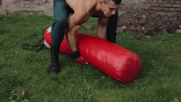 Z bliska. Silny przystojny sportowiec w czerwonych rękawiczkach bokserskich podnosi czerwony worek treningowy, rzucając go na ziemię przez ramię. Zwolniony ruch — Wideo stockowe