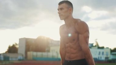 Spor sahasında vücut ağırlığı şınavı yapan üstsüz bir sporcunun yan görüntüsü. Arka planda futbol oynayan insanların bulanık görüntüsü