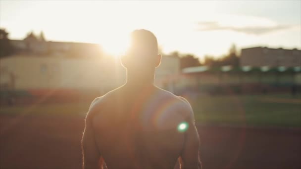 Movimento lento. Retrato de belo jovem sem camisa caminhando em direção à câmera com sol brilhante atrás dele. Vista turva do estádio em segundo plano — Vídeo de Stock