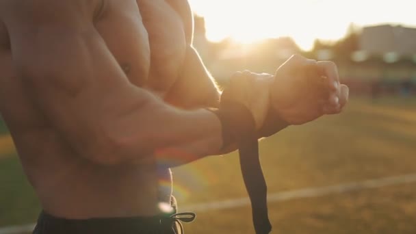 Vista lateral cercana de un deportista musculoso que se pone el soporte de muñeca negro en las manos. Sol brillante en el fondo — Vídeos de Stock