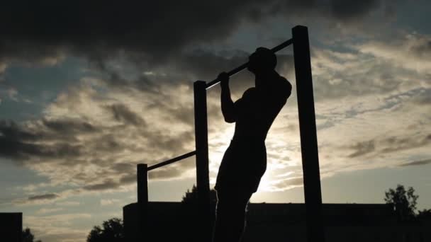 Silueta de un joven fuerte haciendo flexiones en una barra horizontal durante el entrenamiento en la calle. Vista de abajo. Cielo al atardecer en el fondo — Vídeo de stock