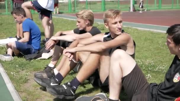 Bobruisk, Bělorusko - 12. srpna 2019: Skupina mladých mužů ve sportovním oblečení sedících na trávě u basketbalového hřiště — Stock video