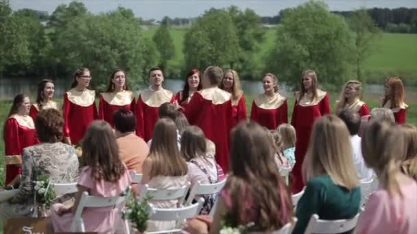 Minsk, Bielorrússia - 24 de agosto de 2019: Coro gospel masculino e feminino em vestes vermelhas cantando com um maestro na frente do público no verão ao ar livre — Vídeo de Stock