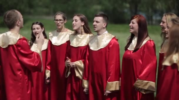 Minsk, Belarus - August 24, 2019: Male and female gospel choir in red robes singing with a conductor in front of the audience in summer outdoors — Stock Video