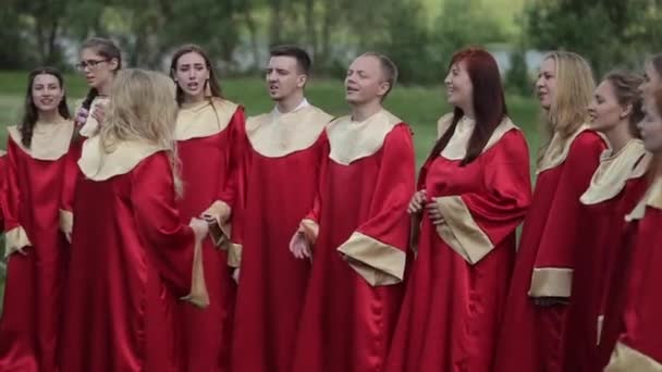 Minsk, Bělorusko - 24. srpna 2019: Sbor mladých mužů a žen v červených kostýmech šťastně zpívá s dirigentem v létě v parku — Stock video