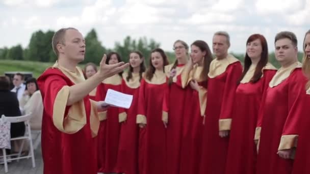 Minsk, Bielorussia - 24 agosto 2019: Veduta posteriore di un direttore d'orchestra che dirige un coro gospel di giovani uomini e donne in abiti rossi in estate all'aperto — Video Stock