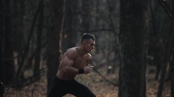 Atleta muscular solitario corre en el bosque entre los árboles con el torso desnudo. Un rodaje cinematográfico. hojas de otoño en el suelo . — Vídeo de stock