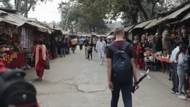 Kathmandu, Nepal - 14 November 2019: A rear back view of a young caucasian male tourist walking through a nepalese market, bazaar. Holding a tripod, camera gear. Turing around, looking at the camera. — Stok video