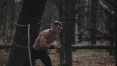 Çıplak gövdeli hızlı koşan kaslı adam. Ormanda genç bir Kafkasyalı koşucu. Ağaçlar arasında görünüm