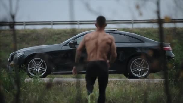 Um homem musculoso está parado à frente de um carro. O conceito de confronto e competição entre carro e homem — Vídeo de Stock