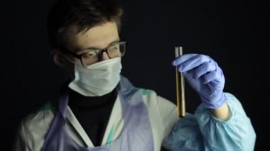 Gözlüklü, tek kullanımlık maske takan beyaz bir doktor. Çözümlü bir ampule bakıyorum. Koyu arkaplan, tıbbi üniforma, kamuflaj.