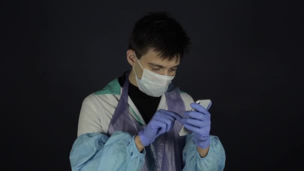 A young caucasian doctor calling someone on the cell phone. Wearing protective medical suit. Isolated dark background. Coronavirus. — Stock Video