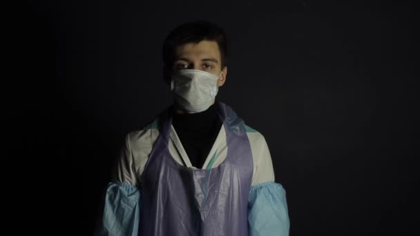 En ung kaukasisk läkare tittar på kameran och visar handflatan med en skylt Stop. Bär medicinsk kostym, handskar, mask. Coronavirus Covid 19. Isolerad på en svart mörk bakgrund. — Stockvideo