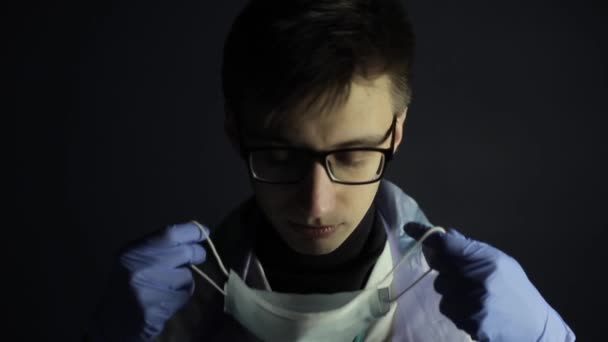 Ein junger kaukasischer Arzt setzt eine medizinische Schutzmaske auf. Daumen hoch. Dunkelschwarzer Hintergrund. Medizinische Handschuhe. — Stockvideo