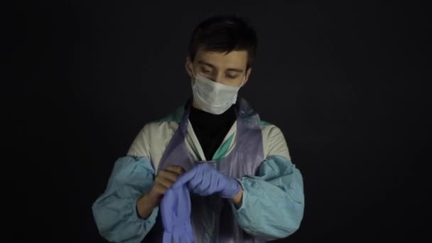 Młody biały mężczyzna zakładający rękawiczki medyczne, patrzący w kamerę. Ubrany w medyczne ubrania ochronne. Odizolowany na czarnym ciemnym tle. Coronawirus Covid19 — Wideo stockowe