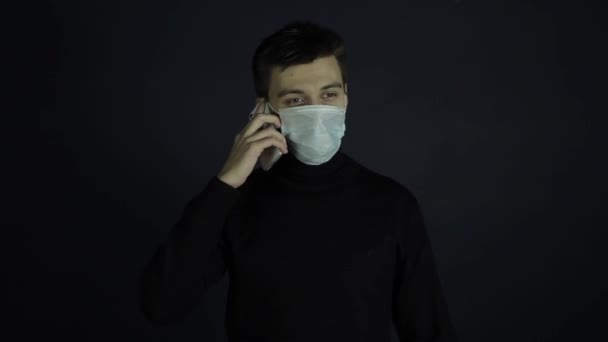 Ένας νεαρός Καυκάσιος μιλάει στο τηλέφωνο φορώντας μια προστατευτική υγιεινή ιατρική μάσκα. Coronavirus, προστασία. Εσωτερικό σκοτεινό φόντο. — Αρχείο Βίντεο