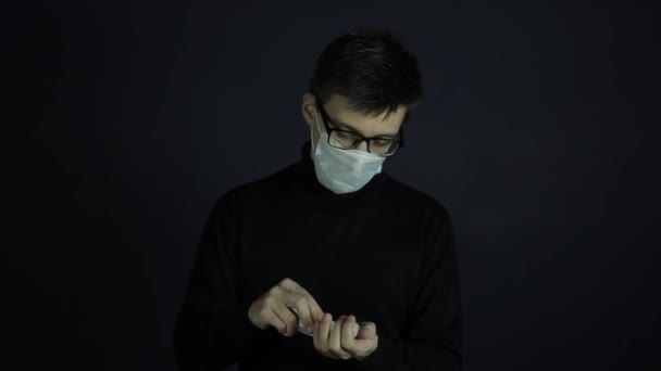 A young caucasian man wearing a hygienic medical mask sanitising his hands. Shot on dark, black background. Looking at the camera. Coronavirus COVID-19 — Stock Video