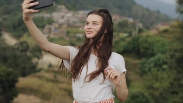 Una joven delgada chica caucásica con el pelo largo y castaño tomando una selfie en la cima de una montaña, una colina en Nepal. Paisaje . — Vídeos de Stock