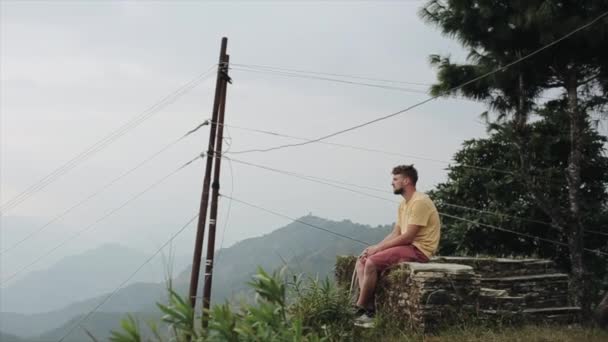 En ung kaukasisk man sitter på toppen av en kulle och njuter av en vacker utsikt över landskapet i Nepal. — Stockvideo