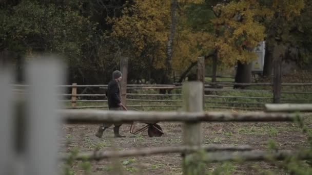 Bobruisk, Vitryssland - 14 oktober 2019: En man går med skottkärra till en boskapsranch längs ett trästaket. Sidovy — Stockvideo
