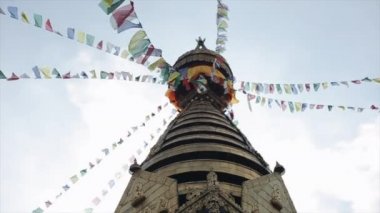 Nepal kulesinin altından bir manzara, dua bayraklı bir tapınak. Gökyüzü açık, güvercinler uçuyor.