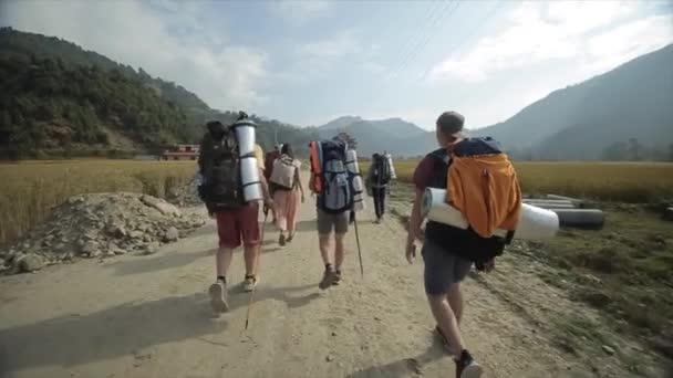 Pokhara, Nepal - 14 November 2019: A back rear view of backpackers walking on a sandy road in Nepal, surrounded by nature, mountains, field. — Stock Video