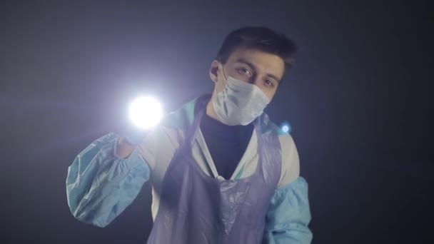 A young caucasian doctor flashing the light at the camera. Studying a COVID-19 coronavirus model. Black dark background. Wearing medical clothes. — Stock Video